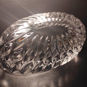 May include: A clear, oval-shaped crystal bowl with a textured, cut-glass design. The bowl's surface reflects light, creating a sparkling effect. The intricate pattern features repeating leaf-like shapes, adding to its elegance.