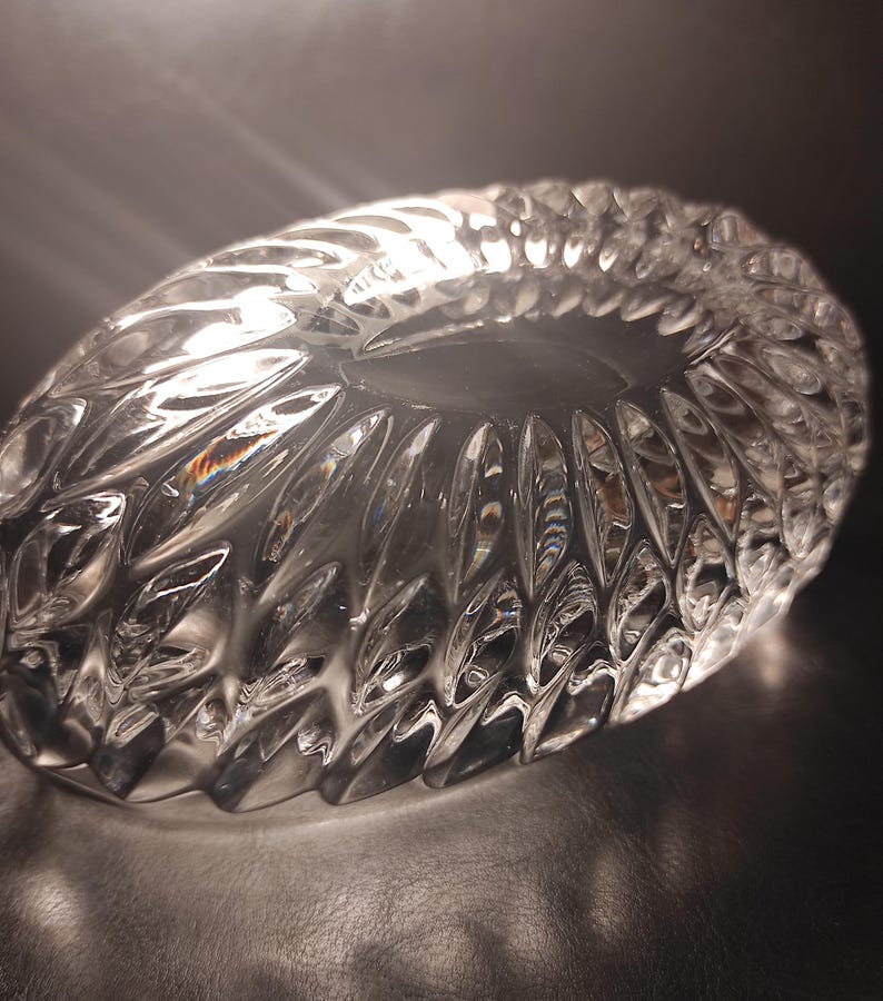 May include: A clear, oval-shaped crystal bowl with a textured, cut-glass design. The bowl's surface reflects light, creating a sparkling effect. The intricate pattern features repeating leaf-like shapes, adding to its elegance.