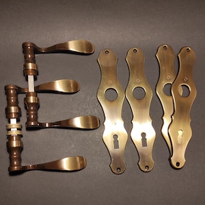 Könnte beinhalten: Ein Set aus vier messingfarbenen Türgriffen mit passenden länglichen Türschildern. Die Griffe haben ein geschwungenes Hebeldesign mit metallischem Finish. Die vier vertikalen Türschilder verfügen über eine runde Öffnung und ein Schlüsselloch.
