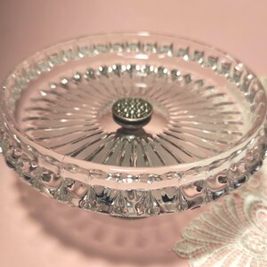 May include: A clear glass pedestal dish featuring a circular tray with a radiating sunburst pattern and a scalloped edge. The centre of the dish is adorned with a small, metallic, circular knob with a textured, dotted surface finish.