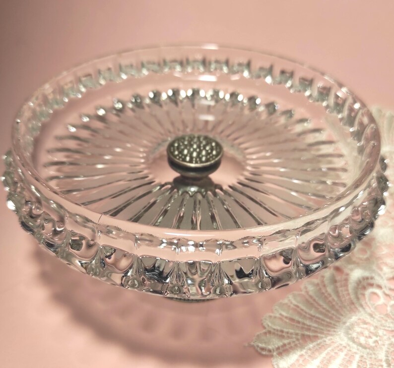 May include: A clear glass pedestal dish featuring a circular tray with a radiating sunburst pattern and a scalloped edge. The centre of the dish is adorned with a small, metallic, circular knob with a textured, dotted surface finish.
