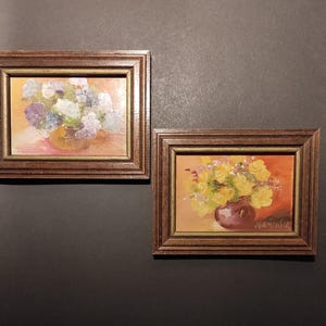 May include: Two framed paintings of floral arrangements in vases. The painting on the left features a bouquet of purple and white flowers in a brown vase. The painting on the right features a bouquet of yellow flowers in a purple vase. The painting on the right is signed "Vannicofer".