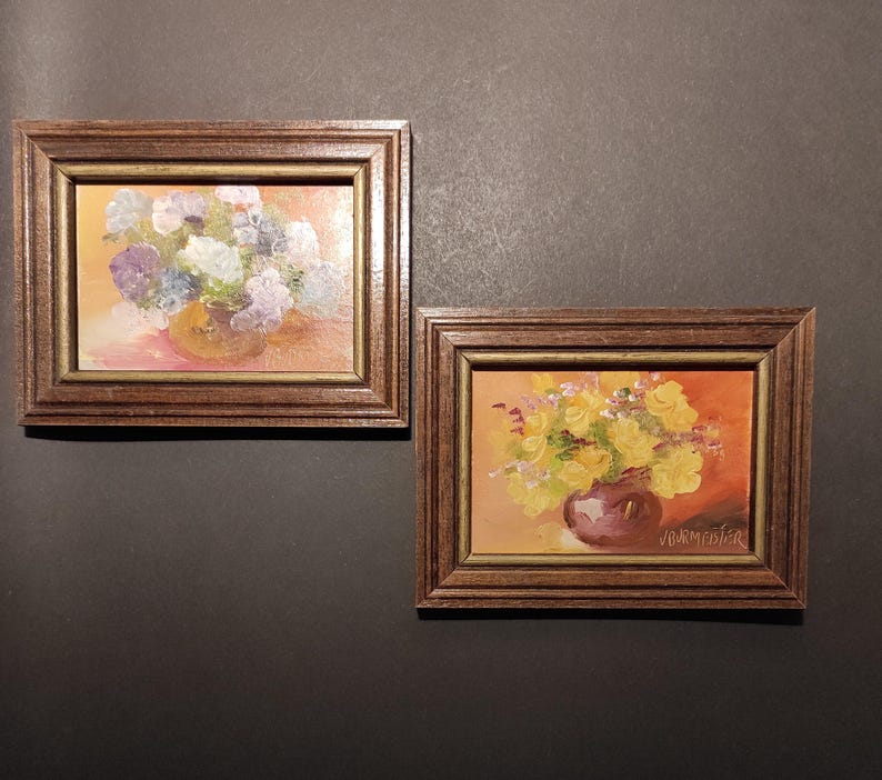 May include: Two framed paintings of floral arrangements in vases. The painting on the left features a bouquet of purple and white flowers in a brown vase. The painting on the right features a bouquet of yellow flowers in a purple vase. The painting on the right is signed "Vannicofer".