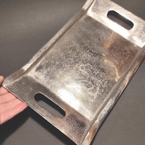 Könnte beinhalten: Ein rechteckiges, silberfarbenes Metalltablett mit einer texturierten, gehämmerten Oberfläche. Das Tablett verfügt über zwei integrierte Griffaussparungen an den kurzen Seiten und einen leicht erhöhten Rand, der eine reflektierende Servierfläche bietet.