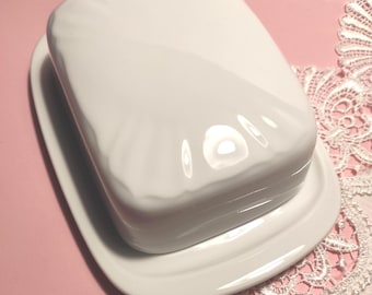 Könnte beinhalten: Eine weiße Butterdose aus Keramik mit rechteckigem Boden und passendem Deckel. Der Deckel hat eine glatte, glänzende Oberfläche mit dezenten, fächerförmigen Prägemustern auf der Oberseite, die ein schlichtes Küchenaccessoire bilden.
