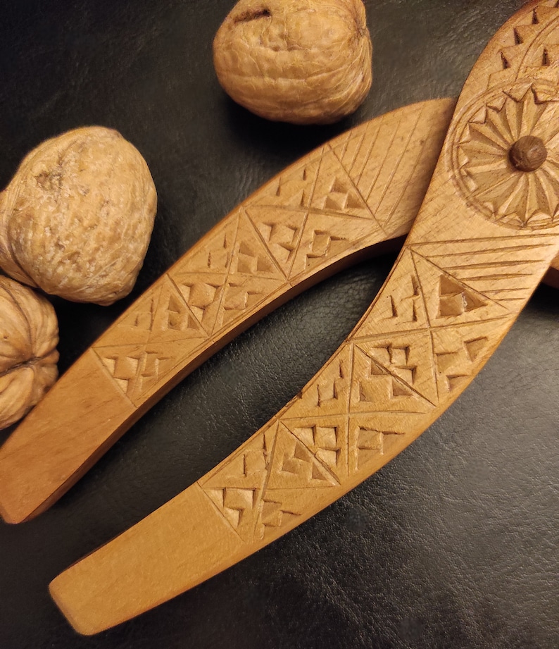 Op de afbeelding: Een houten notenkraker met ingewikkelde houtsnijwerk van geometrische patronen. De notenkraker is gemaakt van lichtbruin hout en heeft een traditioneel ontwerp. Twee walnoten zijn ook afgebeeld.