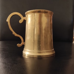 Puede incluir: Una jarra de cerveza de metal dorada con un asa en forma de corazón. La jarra tiene una superficie lisa y pulida.