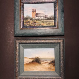 May include: Two framed landscape paintings. The top painting depicts a church with a tall brick tower. The bottom painting shows a sandy beach with dunes and ocean. Both paintings are in dark green frames.