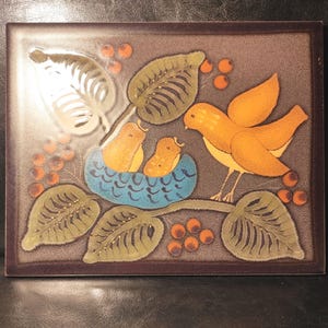 Op de afbeelding: Een rechthoekige keramische tegel met een bruine achtergrond en een getextureerd oppervlak. Het ontwerp toont een oranje vogel op een tak bij een blauw nest met twee kleine gele kuikens, omringd door groene bladeren en oranje bessen.