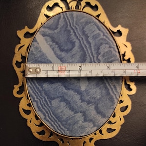 Puede incluir: Un marco de fotos ovalado dorado y ornamentado con un respaldo de tela azul. El marco mide aproximadamente 10 cm de ancho. Las palabras "MADE IN ITALY" son visibles. El marco est&aacute; sobre una superficie negra.