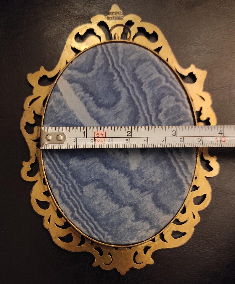 Puede incluir: Un marco de fotos ovalado dorado y ornamentado con un respaldo de tela azul. El marco mide aproximadamente 10 cm de ancho. Las palabras "MADE IN ITALY" son visibles. El marco est&aacute; sobre una superficie negra.