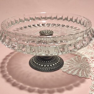 May include: A clear glass pedestal dish featuring a scalloped rim and a radial pattern on the interior surface. The dish is supported by a dark, metallic, circular base with a textured, beaded design around the perimeter and a central stud.