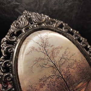 May include: An oval-shaped picture frame with an ornate, silver-colored metal frame. The frame contains a vintage-style print of a tree with brown branches and a light brown background.