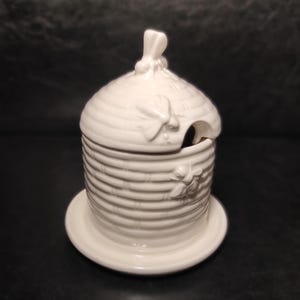 May include: A white ceramic honey pot with a lid and a small spoon. The pot is shaped like a beehive with horizontal ridges and bee-shaped embellishments. It sits on a matching saucer.
