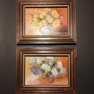 May include: Two framed oil paintings of floral arrangements. The top painting features yellow flowers in a vase with a warm orange background. The bottom painting features purple and white flowers in a vase with a pink background. Both paintings are signed by the artist.