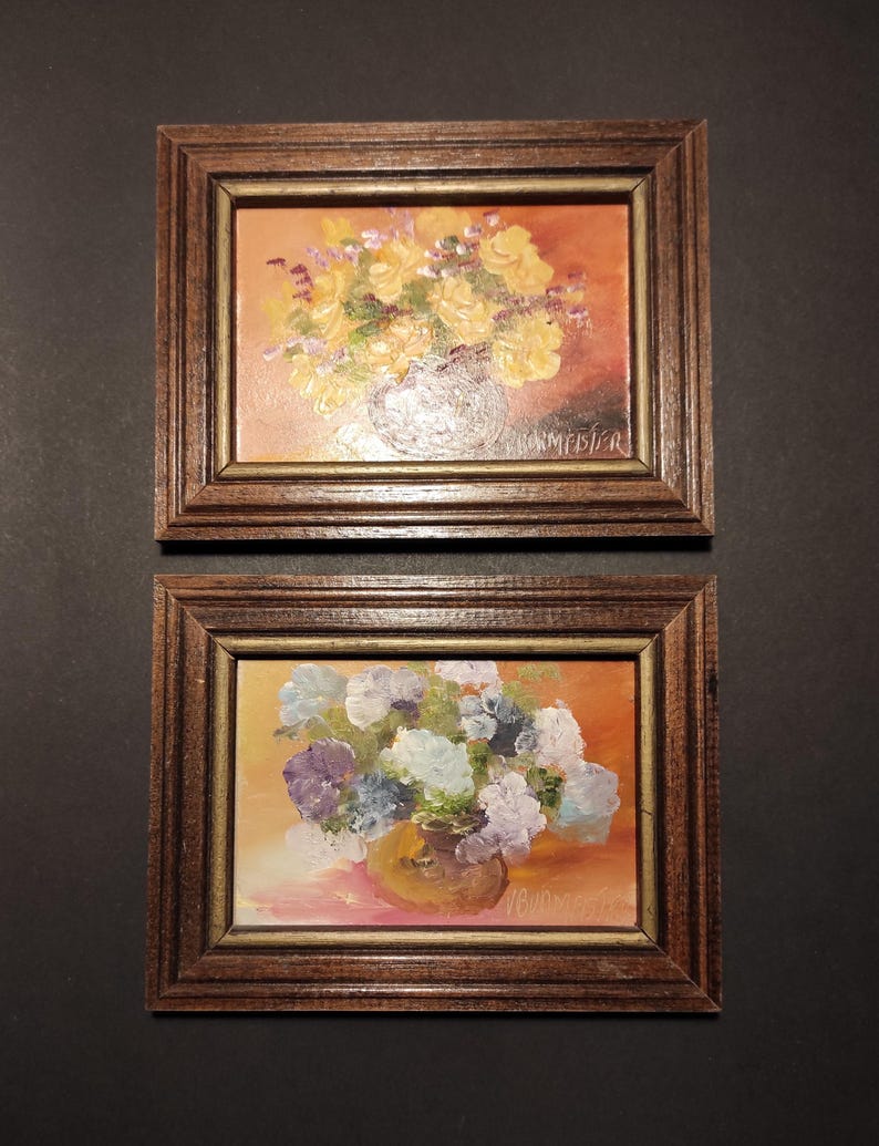 May include: Two framed oil paintings of floral arrangements. The top painting features yellow flowers in a vase with a warm orange background. The bottom painting features purple and white flowers in a vase with a pink background. Both paintings are signed by the artist.