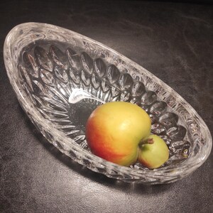 May include: A clear, oval-shaped crystal bowl with a textured, cut-glass design. Inside the bowl are two apples, one yellow and red, the other green. The bowl sits on a dark surface.