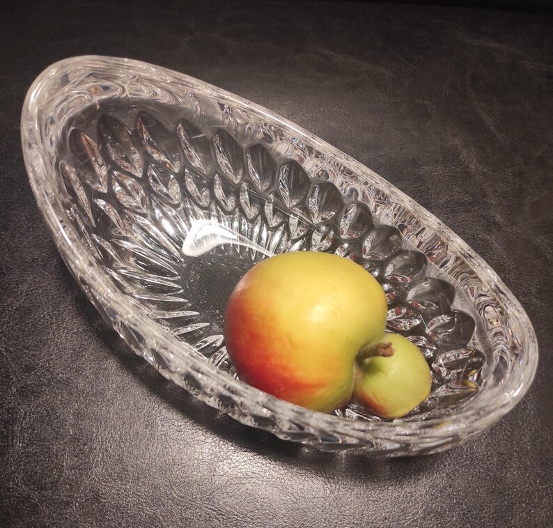 May include: A clear, oval-shaped crystal bowl with a textured, cut-glass design. Inside the bowl are two apples, one yellow and red, the other green. The bowl sits on a dark surface.