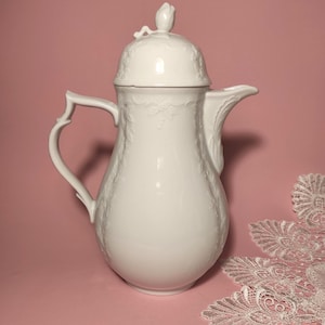 Op de afbeelding: Een witte porseleinen koffiepot met een deksel en een handvat. De pot heeft een bloemenmotief en een tuit. Het deksel is bekroond met een decoratieve bloem. De pot staat op een roze achtergrond.