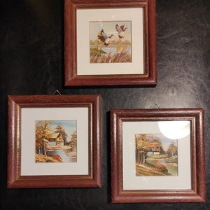 以下が含まれることがあります： 茶色の木製フレームに入った3つの風景画。絵画は、飛んでいるアヒル、川沿いの家、秋の木々など、自然のシーンを描いています。アートワークは白いマットで額装されています。