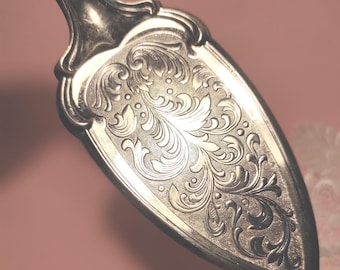 Könnte beinhalten: Ein Tortenheber aus silberfarbenem Metall mit einer spitz zulaufenden, dreieckigen Klinge, die mit einem komplexen, gravierten Rankenmuster verziert ist. Der Griff geht in einen glatten, polierten Hals über, der zur strukturierten Klinge führt.