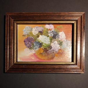 May include: A framed oil painting depicting a bouquet of purple, white and blue flowers in a brown vase. The flowers are set against a warm orange background.
