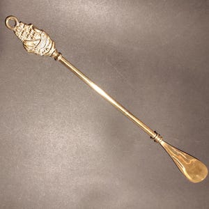 May include: A gold metal shoehorn with a decorative ship design at the top.