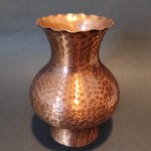 Könnte beinhalten: Eine Kupfervase mit gehämmertem Muster und einem ausgestellten, gewellten Rand. Die Vase hat einen bauchigen Körper und einen kleinen, kreisförmigen Sockel. Das Kupfer hat einen warmen, rötlich-braunen Farbton.
