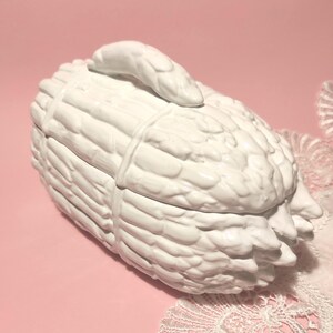 Op de afbeelding: Witte keramische sieradendoos in de vorm van een zwaan, met een textuur van veren. Het deksel heeft een zwanenkop. De doos staat op een roze ondergrond met kanten rand.