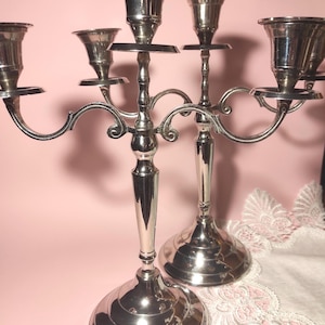 Può includere: Due candelabri d'argento con più bracci, ciascuno con una coppa per candele. I candelabri hanno un design decorativo e curvo e poggiano su una base rotonda. Lo sfondo è rosa tenue.