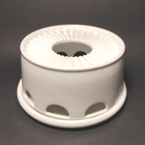 Może przedstawiać: Biały ceramiczny podgrzewacz do herbaty o okrągłym kształcie. Podgrzewacz ma okrągłą podstawę i cylindryczny korpus z łukowatymi wycięciami. Górna część ma pofałdowaną krawędź wokół centralnego otworu. Minimalistyczny i funkcjonalny design.