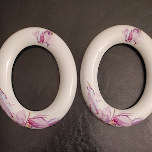 May include: Two white ceramic ring holders with a floral design. The design features pink and purple flowers with a watercolor effect.