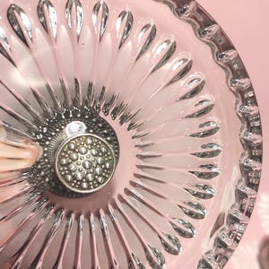 May include: A clear glass dish featuring a radial pattern of elongated, petal-like indentations extending from the centre. The centre of the dish is adorned with a circular, metallic silver-toned knob that has a textured, dotted surface design.