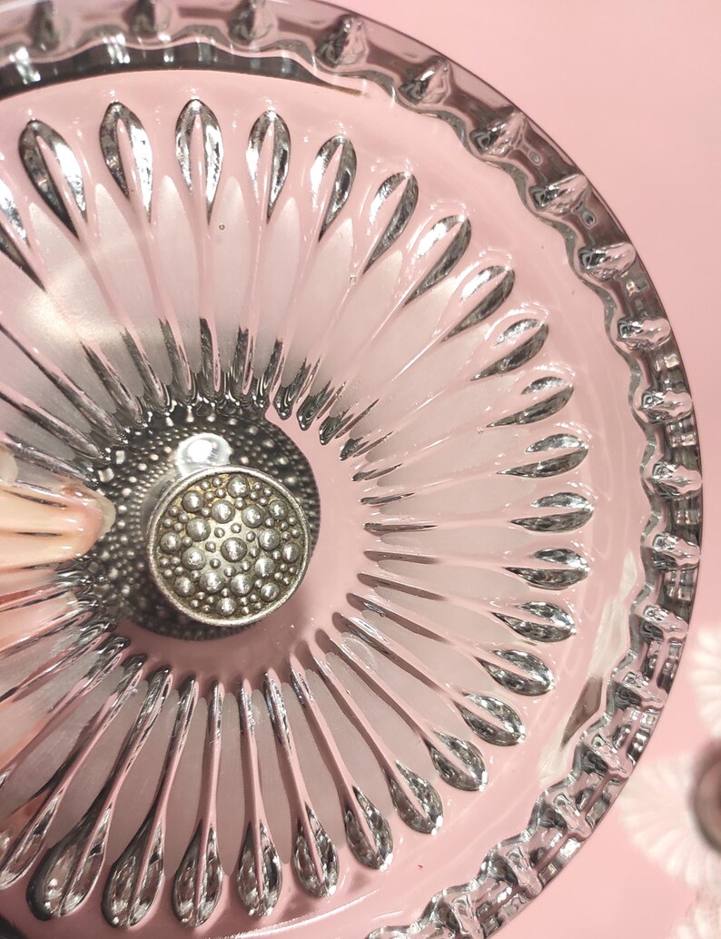 May include: A clear glass dish featuring a radial pattern of elongated, petal-like indentations extending from the centre. The centre of the dish is adorned with a circular, metallic silver-toned knob that has a textured, dotted surface design.