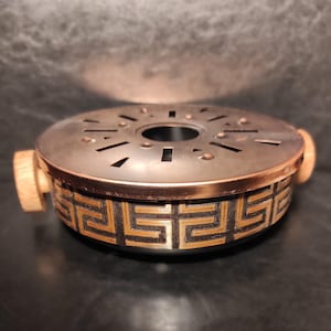 Könnte beinhalten: Ein runder Räuchergefäß aus dunklem Metall mit Kupferrand und Holzgriffen. Der Sockel hat ein geometrisches Gold- und Schwarzmuster. Die Oberseite hat ausgeschnittene Formen und ein zentrales Loch.