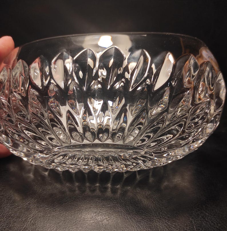 May include: A clear, cut-glass bowl with a detailed, repeating leaf-like pattern. The bowl has a rounded shape and a textured exterior, reflecting light. The design is symmetrical, creating a decorative and elegant appearance. The bowl is likely used for serving or display.