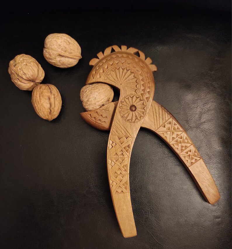 Op de afbeelding: Een houten notenkraker met ingewikkelde houtsnijwerk in de vorm van een vogel. De notenkraker is open, met een walnoot erin. Er liggen drie walnoten op de tafel.