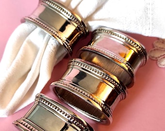 Op de afbeelding: Een set van vier gepolijste zilverkleurige metalen servetringen met een gladde centrale band en kralendetails langs de boven- en onderrand. Eén ring bevat een witte stoffen servet met een delicaat kantachtig patroon langs de rand.