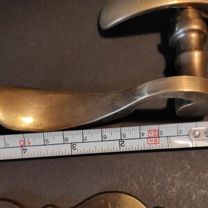 May include: A vintage brass door handle with a curved lever and a decorative backplate. The handle measures approximately 4 inches in length. The brass has a warm, aged patina, suggesting its antique nature. The handle is likely for a door or cabinet.