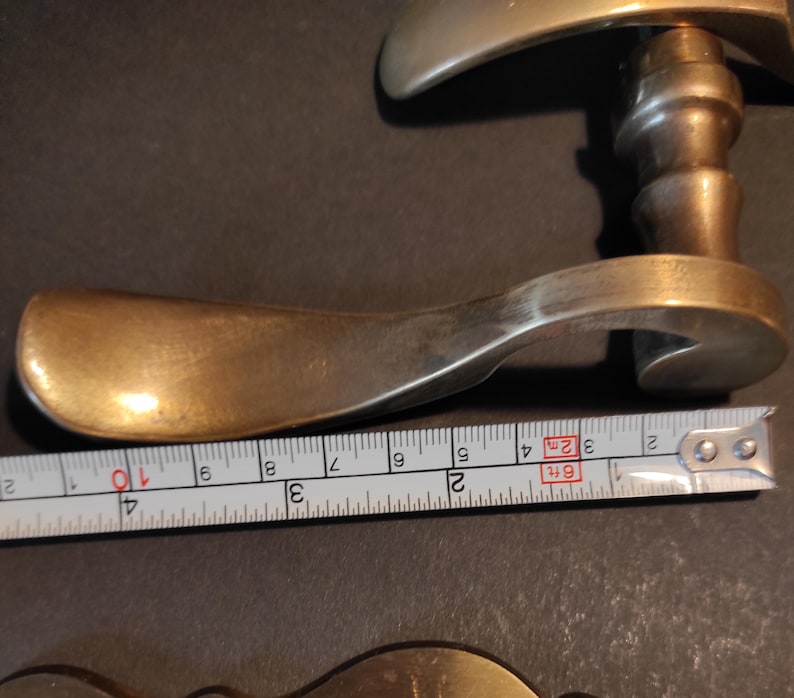 May include: A vintage brass door handle with a curved lever and a decorative backplate. The handle measures approximately 4 inches in length. The brass has a warm, aged patina, suggesting its antique nature. The handle is likely for a door or cabinet.