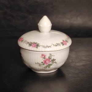 May include: White ceramic sugar bowl with a lid, decorated with pink and green rose floral patterns. The bowl has vertical ribbed detailing and a conical finial. A decorative item for the kitchen or dining room.