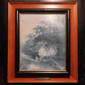 Op de afbeelding: Een ingelijst schilderij van een landelijk tafereel met een huis in de verte, bomen en een pad dat naar het huis leidt. Het schilderij is in tinten blauw en wit, en de lijst is bruin met een zwarte binnenrand.