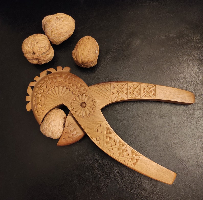 Op de afbeelding: Een houten notenkraker met ingewikkelde houtsnijwerk en een decoratief ontwerp. De notenkraker is open en heeft een noot erin. Er liggen vier walnoten op de tafel naast de notenkraker.
