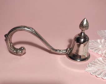 Könnte beinhalten: Ein silberfarbener Kerzenlöscher aus Metall mit einem geschwungenen, strukturierten Griff und einem glockenförmigen Kopf, der von einer kleinen, spitzen Verzierung gekrönt wird. Der Griff weist ein verziertes, geprägtes Muster auf.
