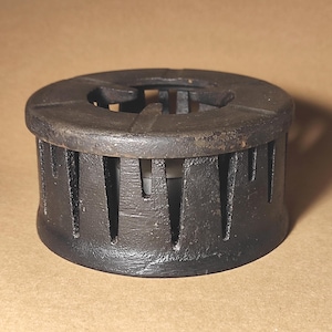 Puede incluir: Un salvamanteles oscuro de hierro fundido con un diseño circular. El salvamanteles tiene ranuras verticales en los lados y una parte superior estampada. Diseñado para proteger las superficies del calor.