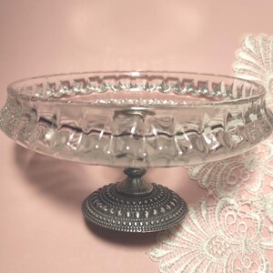 May include: A clear glass bowl with a textured, scalloped rim, supported by a circular, dark metallic pedestal base featuring a detailed, embossed pattern. The bowl has a shallow, wide design and sits atop a sturdy, ornate metal stand.
