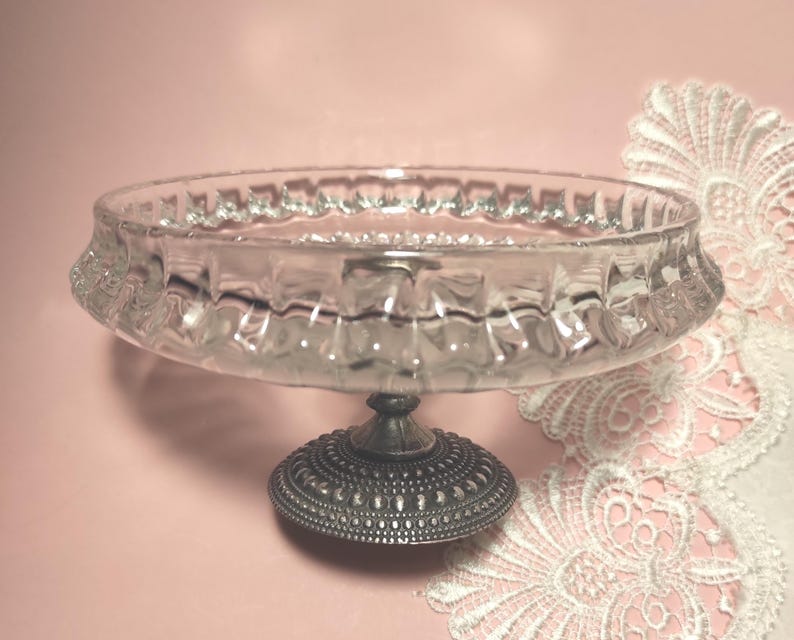 May include: A clear glass bowl with a textured, scalloped rim, supported by a circular, dark metallic pedestal base featuring a detailed, embossed pattern. The bowl has a shallow, wide design and sits atop a sturdy, ornate metal stand.