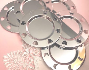 Puede incluir: Una colección de platos plateados brillantes. Cada plato tiene un diseño circular con recortes en forma de corazón alrededor del borde. Los platos están dispuestos sobre una superficie rosa con un detalle de encaje blanco.