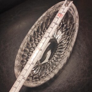 May include: Clear, oval-shaped glass dish with a textured, cut-glass design. A measuring tape is laid across the dish, showing a length of approximately 7 inches. The dish is likely for serving or display.