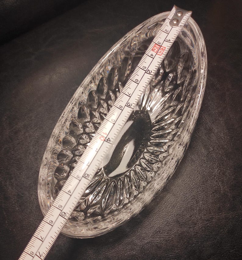 May include: Clear, oval-shaped glass dish with a textured, cut-glass design. A measuring tape is laid across the dish, showing a length of approximately 7 inches. The dish is likely for serving or display.